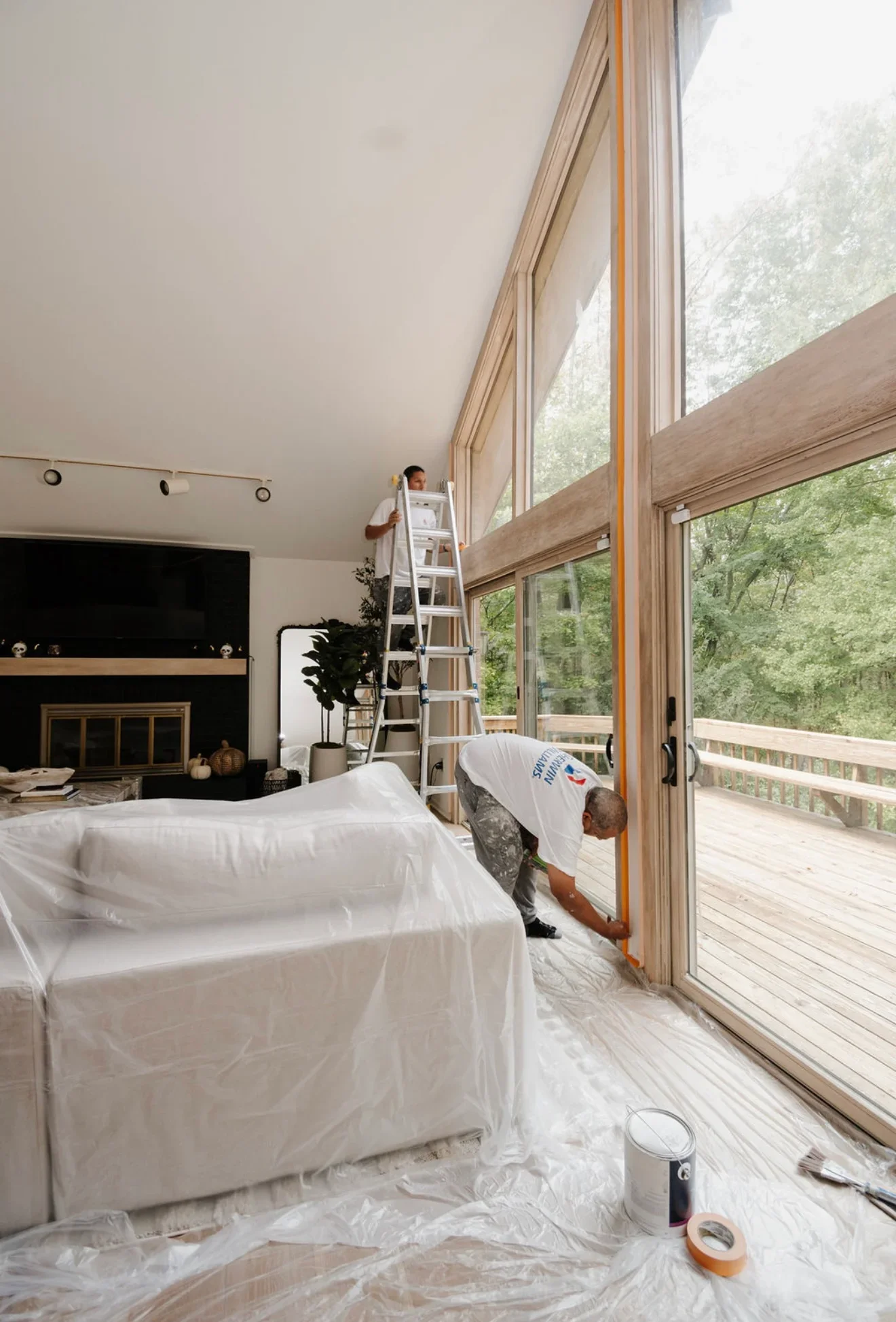Interior painting in progress with white ceiling paint, protective sheeting, and ladder setup in bedroom with glass doors